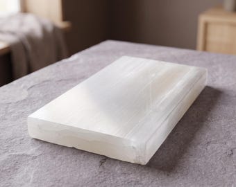May include: A rectangular slab of white selenite crystal with a smooth, polished top surface and a fibrous, translucent appearance. The stone features natural linear striations and a glossy finish, showcasing its crystalline structure and white colour.