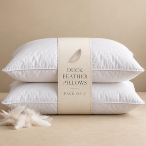 May include: Two white rectangular pillows stacked vertically, wrapped in a beige paper band featuring a feather illustration and the text DUCK FEATHER PILLOWS PACK OF 2. A small cluster of loose white feathers sits in the foreground.