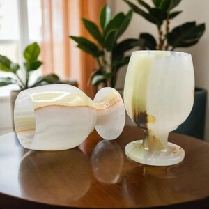 May include: Two onyx wine glasses and a matching napkin ring sit on a wooden table. The glasses have a rounded bowl and a short stem. The napkin ring is a rounded rectangle. The onyx is white with bands of yellow, brown, and green.