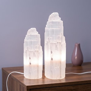 May include: Two white selenite crystal lamps with a jagged, natural tower-like structure. Each lamp features a cylindrical base and emits a soft, warm internal glow from the bottom, highlighting the vertical striations and translucent texture.