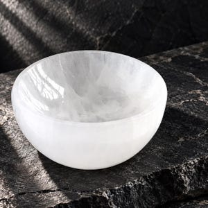 May include: A round, translucent white selenite bowl with a smooth, polished interior surface and a natural, crystalline texture. The bowl features a curved, hemispherical shape with a consistent white colour throughout its solid mineral structure.