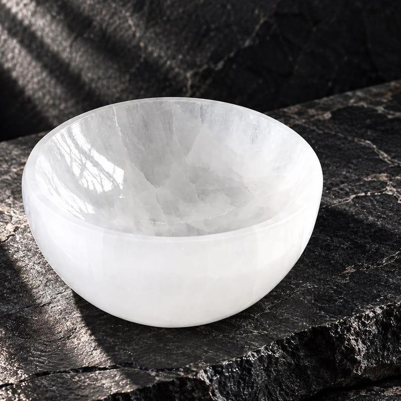 May include: A round, translucent white selenite bowl with a smooth, polished interior surface and a natural, crystalline texture. The bowl features a curved, hemispherical shape with a consistent white colour throughout its solid mineral structure.