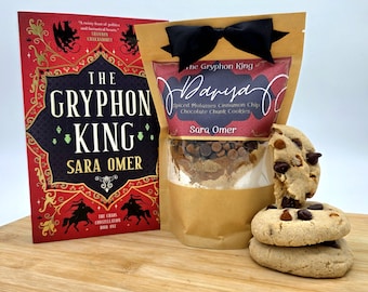 May include: A book titled "The Gryphon King" next to a bag of cookie mix and a stack of cookies. The book has a red cover with black text and illustrations. The cookie mix is in a clear bag with a brown paper wrapper and a black bow.