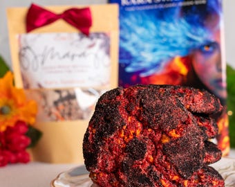 Pode incluir: Uma pilha de biscoitos redondos e texturizados com um interior vermelho vibrante e um revestimento exterior escuro de aspeto carbonizado, servidos num prato branco com rebordo dourado. Ao fundo, um livro de Kara Swanson e uma bolsa bege.