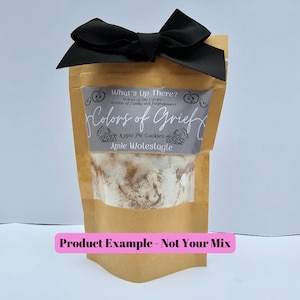 May include: A brown paper bag with a black ribbon tied around the top. The bag contains a mix of ingredients for apple pie cookies. The bag has a label that says "What's Up There? Voices of the Future: Stories of Family and Fearlessness Colors of Grief Apple Pie Cookies Amie Woleslagle". The text "Product Example - Not Your Mix" is written in pink below the bag.