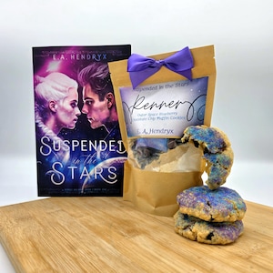 May include: A brown paper bag with a purple ribbon tied around it. The bag has a label that says "Suspended in the Stars Renner Outer Space Blueberry Chocolate Chip Muffin Cookies E.A. Hendryx". There is a book titled "Suspended in the Stars" by E.A. Hendryx in the background. Three blueberry muffin cookies are on a wooden surface in front of the bag.