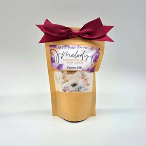 May include: A kraft paper bag filled with cookies, tied with a burgundy bow. The bag has a clear window showing the cookies inside. The label reads "All for Mage and Melody" and "Melody Espresso Cheesecake M&M Cookies Emma Hill."