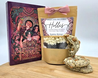 May include: A book titled "Down with the Prince" next to a bag of "Hollis" Caramel Black Tea White Chocolate Chip Cookies. The cookie bag has a pink bow. Two cookies are stacked in front of the bag. The book and bag are on a wooden cutting board.