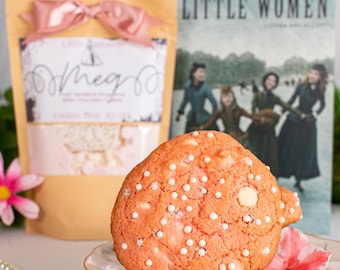 May include: A close-up of a pink cookie with white chocolate chips on a decorative plate, next to a book titled "Little Women" and a bag with a pink bow. A pink flower and a string of pearls are also visible.