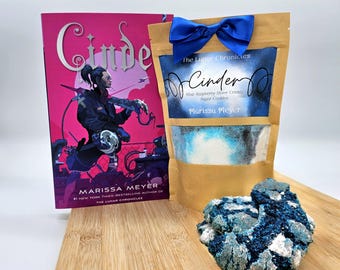 May include: A brown paper bag with a blue ribbon tied around it. The bag has a clear window showing blue and white sugar cookie mix inside. The bag is labeled "The Lunar Chronicles Cinder Blue Raspberry Silver Crinkle Sugar Cookies Marissa Meyer". A paperback book titled "Cinder" by Marissa Meyer is next to the bag. A blue and white sugar cookie is in the foreground.