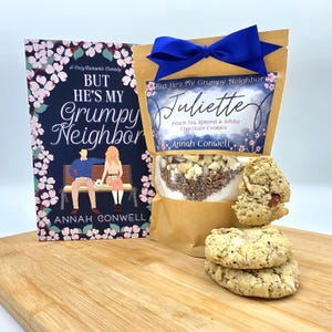 May include: A brown paper bag with a blue ribbon tied around it. The bag has a label that says "But He's My Grumpy Neighbor Juliette Peach Tea Almond & White Chocolate Cookies Annah Conwell". There is a book with a pink and blue cover that says "But He's My Grumpy Neighbor" and a stack of cookies.
