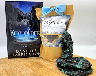 May include: A brown paper bag with a blue ribbon tied around it. The bag is labeled "Keyholder Octavia" and contains blue raspberry rice crispy cheesecake black crinkle cookies. A book titled "Keyholder" by Danielle Harrington is also pictured.