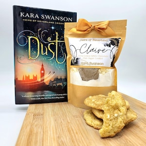 May include: A brown paper bag with a white label that says "Heirs of Neverland Claire Zesty Lemon White Chocolate Pixie Sugar Cookies Kara Swanson". The bag is tied with a gold ribbon. A book titled "Dust" by Kara Swanson is in the background. There are also cookies on a wooden surface.