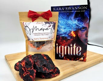 May include: A brown paper bag with a red bow, labeled "IGNITE Mana Cracked Molten Red Velvet Cinnamon Chip Cookies Kara Swanson". The bag contains red and orange sprinkles. A book titled "KARA SWANSON PHOENIX FLAME BOOK ONE" with the word "ignite" in large letters is next to the bag. There are three red and black cookies on a wooden cutting board.