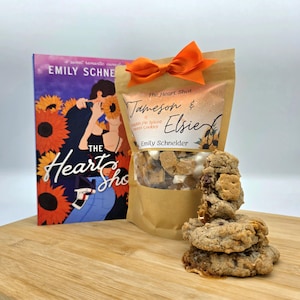 May include: A brown paper bag with a bow tied around the top. The bag contains pumpkin spice cookies and has a label that reads "The Heart Shot Jameson & Elsie Pumpkin Pie Spiced Sweet Cookies Emily Schneider". A book titled "The Heart Shot" by Emily Schneider is in the background. There are three cookies on a wooden surface.