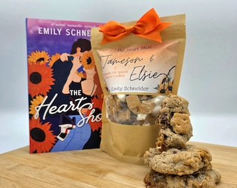 May include: A brown paper bag with a bow tied around the top. The bag contains pumpkin spice cookies and has a label that reads "The Heart Shot Jameson & Elsie Pumpkin Pie Spiced Sweet Cookies Emily Schneider". A book titled "The Heart Shot" by Emily Schneider is in the background. There are three cookies on a wooden surface.