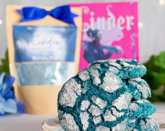 Pode incluir: Uma pilha de quatro biscoitos craquelados redondos de cor azul-petróleo, cobertos com uma camada espessa de açúcar prateado cintilante. Os biscoitos estão dispostos num prato branco, com uma bolsa bege com um laço azul e um livro rosa vivo.
