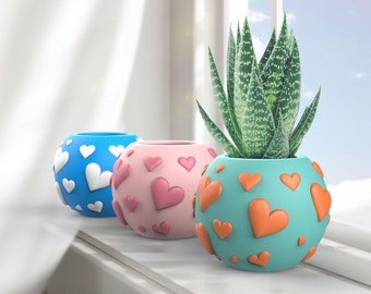 May include: Three ceramic plant pots with heart patterns. The pots are blue, pink, and turquoise. The turquoise pot has a succulent plant inside.