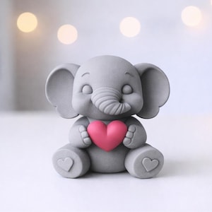 Può includere: Una statuina di elefante grigio che tiene un cuore rosa. L'elefante ha grandi orecchie, una proboscide arricciata e un'espressione sorridente. Ci sono anche cuori sui piedi dell'elefante. La statuina è su una superficie bianca.