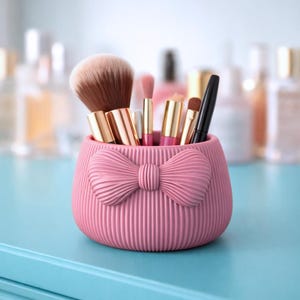 May include: A pink makeup brush holder with a bow design, holding various makeup brushes. The holder has a ribbed texture and is sitting on a blue surface. The brushes include a large powder brush and several smaller eye and lip brushes.