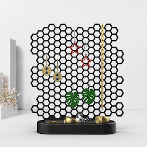 May include: Black metal jewelry stand with a honeycomb design. The stand has a tray for rings and a vertical section for earrings. The earrings include star-shaped, round, and leaf-shaped designs.