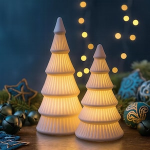 Könnte beinhalten: Zwei weiße Weihnachtsbaumdekorationen aus Keramik mit geripptem Design. Die Bäume sind von innen beleuchtet und erzeugen ein warmes Leuchten. Der Hintergrund umfasst Weihnachtsschmuck und Lichter.