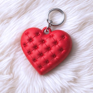 Könnte beinhalten: Ein roter herzförmiger Schlüsselanhänger mit einem gesteppten Design und einem silbernen Schlüsselring. Der Schlüsselanhänger besteht aus einem weichen Material und befindet sich vor einem weißen, flauschigen Hintergrund.