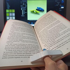 A white plastic thumb book page holder holds open an open book on a wooden surface.
