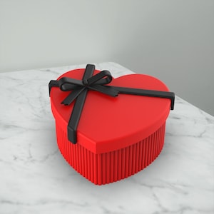 May include: A red heart-shaped gift box with a black ribbon and bow. The box is sitting on a white marble surface.