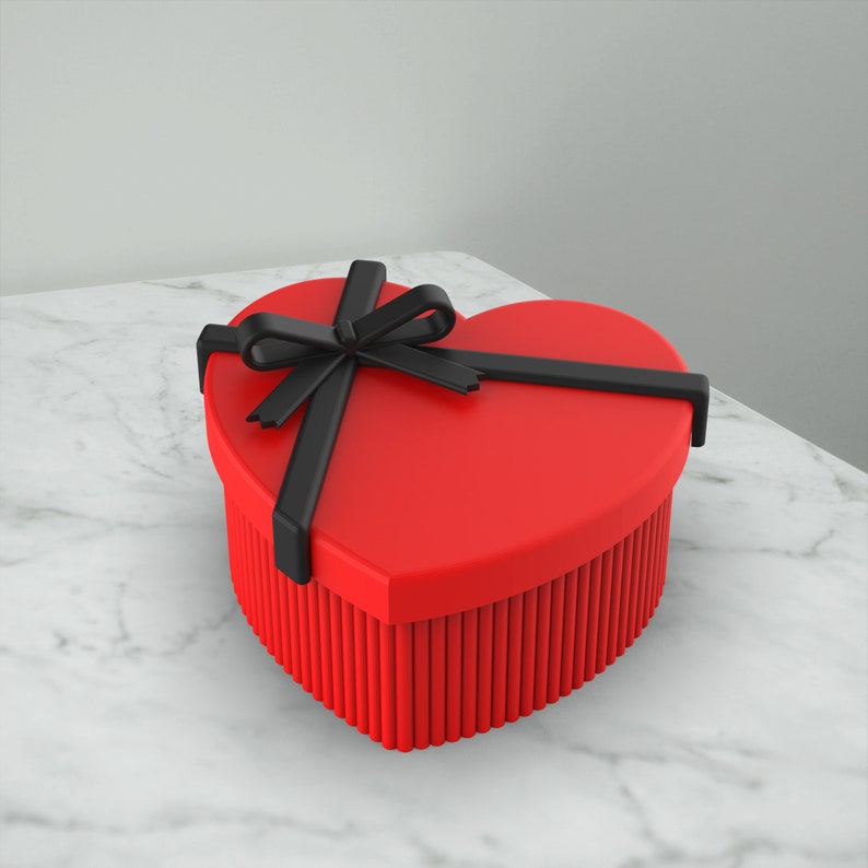 May include: A red heart-shaped gift box with a black ribbon and bow. The box is sitting on a white marble surface.