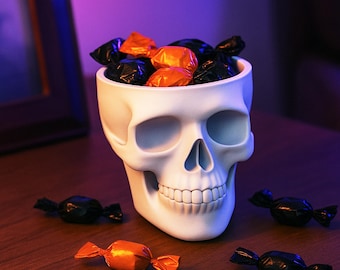 May include: A white ceramic skull-shaped candy bowl filled with orange and black wrapped candies. Several candies are scattered around the base of the bowl. The Halloween-themed bowl has a detailed facial structure.