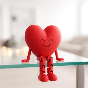 May include: A red heart-shaped ornament with a smiling face, arms, and jointed legs. The figurine is sitting on a glass surface. The heart has a textured surface and is a bright, solid red colour. The arms and legs are also red.