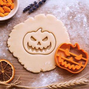 May include: A pumpkin-shaped cookie cutter next to a cut-out pumpkin design in dough. The orange cutter is on a wooden surface with flour, seeds, and dried orange slices. A Halloween-themed baking concept.