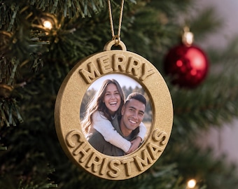 May include: A circular gold-toned metal Christmas ornament featuring a central photograph of a smiling couple. The frame has the raised text MERRY CHRISTMAS embossed around the perimeter and includes a gold hanging string at the top.