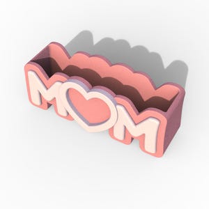 K&ouml;nnte beinhalten: Ein rosa und wei&szlig;er MOM-Pflanzer mit einem Herz in der Mitte. Der Pflanzer ist aus einem weichen, strukturierten Material gefertigt und hat einen gewellten Rand.