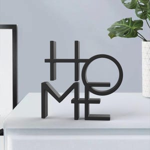May include: Black metal "HOME" decorative sculpture. The letters are stylized and connected, with the "O" forming a circle. The sculpture is placed on a white surface, with a framed picture and a potted plant in the background.