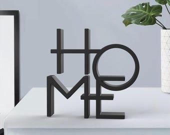 Könnte beinhalten: Schwarze Metallskulptur "HOME". Die Buchstaben sind stilisiert und verbunden, wobei das "O" einen Kreis bildet. Die Skulptur steht auf einer weißen Oberfläche, im Hintergrund ein gerahmtes Bild und eine Topfpflanze.