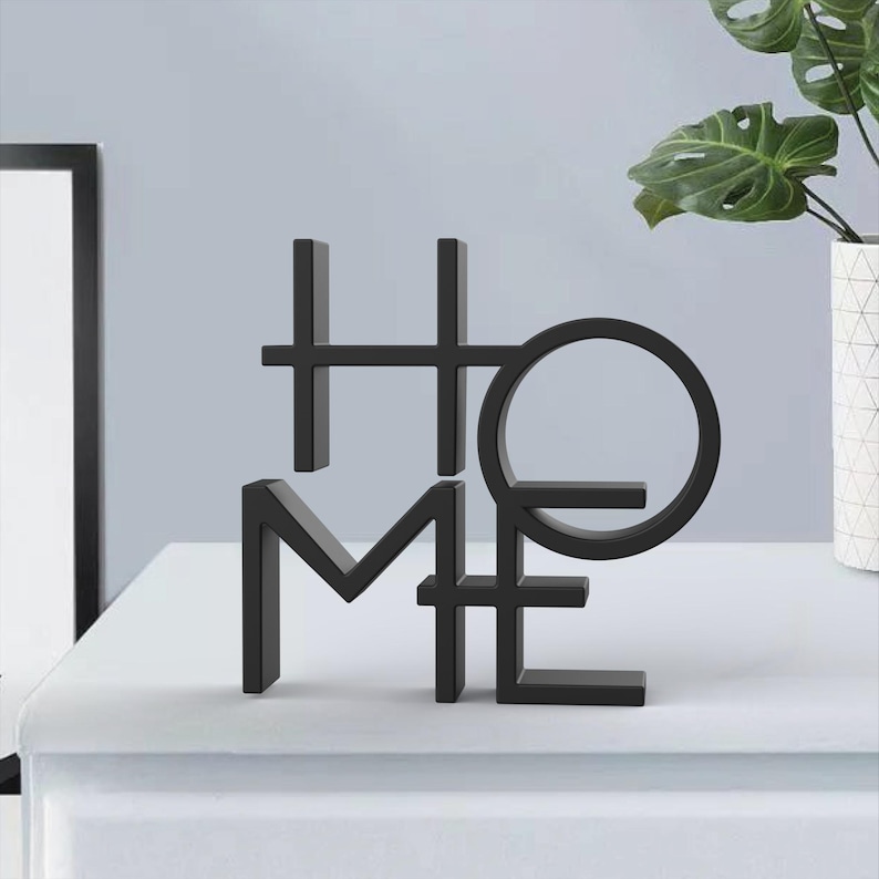 May include: Black metal "HOME" sculpture. The letters are stylized and connected, with the "O" forming a complete circle. The sculpture is placed on a white surface, with a framed picture and a vase with a plant in the background.