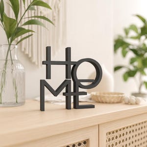 May include: A black metal sculpture spelling out the word "HOME" in a modern, geometric font. The sculpture is displayed on a light-colored wooden surface, with greenery and decorative items in the background.