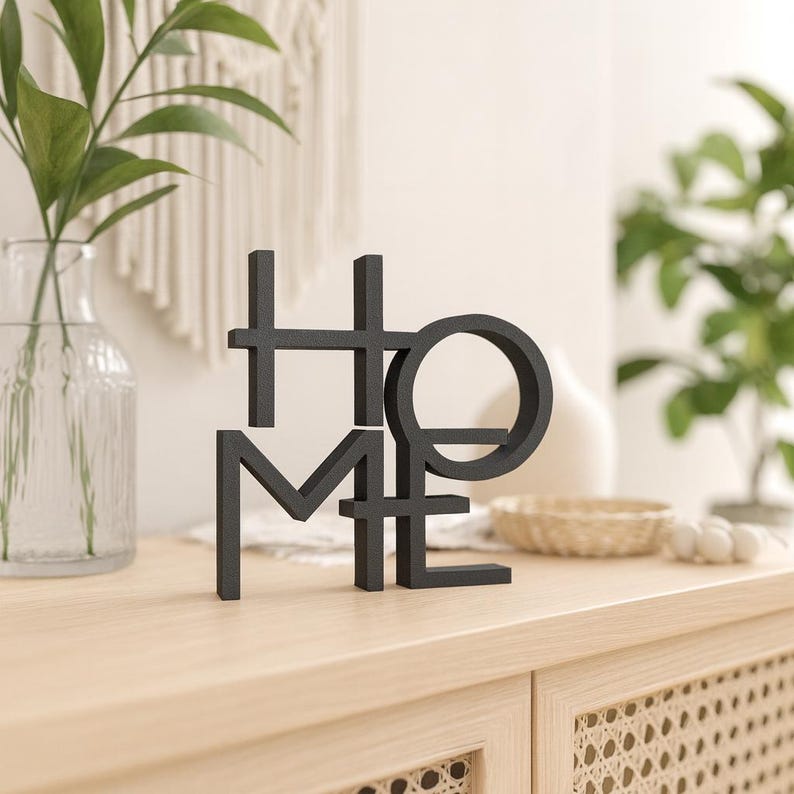 May include: A black metal sculpture spelling out the word "HOME" in a modern, geometric font. The sculpture is displayed on a light-colored wooden surface, with greenery and decorative items in the background.