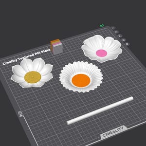 Op de afbeelding: Een digitale interface toont een Creality Textured PEI-plaat met drie witte bloemvormige schalen met gele, oranje en roze centra, samen met een witte cilindrische staaf en een klein veelkleurig kubusblok op een rasterindeling.