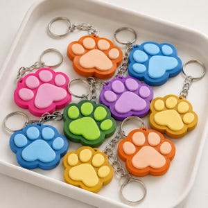 May include: A collection of nine paw print keychains in various colours including orange, blue, pink, purple, green, and yellow. Each keychain features a metal ring and chain attached to a two-toned silicone paw print charm with raised pads.