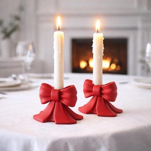 Puede incluir: Dos velas blancas encendidas en candelabros rojos con forma de lazo sobre un mantel blanco. Los candelabros tienen un diseño de lazo detallado. El fondo incluye una chimenea y un comedor.