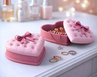 Puede incluir: Cajas de joyería rosas en forma de corazón con un diseño acolchado y detalles de lazo. Una caja está abierta, revelando collares y otras joyas. Varios anillos se exhiben cerca. Frascos de perfume en el fondo.