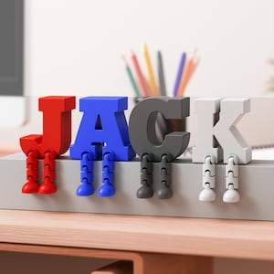 May include: A colorful desk accessory spelling out the name "JACK". The letters are red, blue, gray, and white, each with small, articulated legs. The letters are sitting on a gray surface.