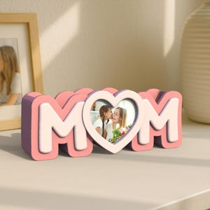 K&ouml;nnte beinhalten: Rosa-wei&szlig;er Holzbildrahmen in Form des Wortes "MOM" mit einem herzf&ouml;rmigen Ausschnitt in der Mitte. Der herzf&ouml;rmige Ausschnitt enth&auml;lt ein Foto von zwei Personen.