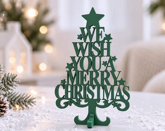 Puede incluir: Una decoración de árbol de Navidad de madera cortada en color verde oscuro, con una estrella en la parte superior y el texto "WE WISH YOU A MERRY CHRISTMAS" integrado. El árbol se sostiene sobre una base decorativa con pies curvos.