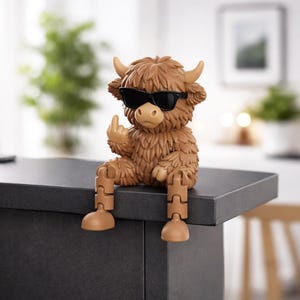 May include: A brown figurine of a Highland cow wearing black sunglasses, giving the middle finger. The cow has articulated legs and is sitting on a dark gray surface. The figurine has a shaggy coat and small horns.