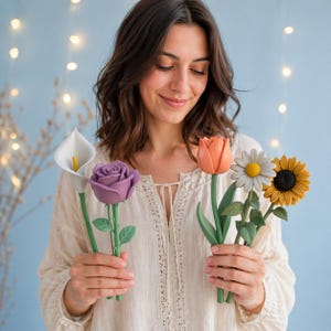 May include: A collection of five artificial flower pens featuring a white calla lily, a purple rose, an orange tulip, a white daisy with a yellow center, and a yellow sunflower, each with a green stem and leaves, designed as functional writing tools.