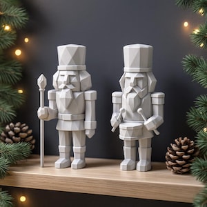 May include: Two gray, geometric-style nutcracker figurines, one holding a staff and the other a drum with drumsticks. The figures are on a wooden shelf, with pinecones and evergreen branches in the background.